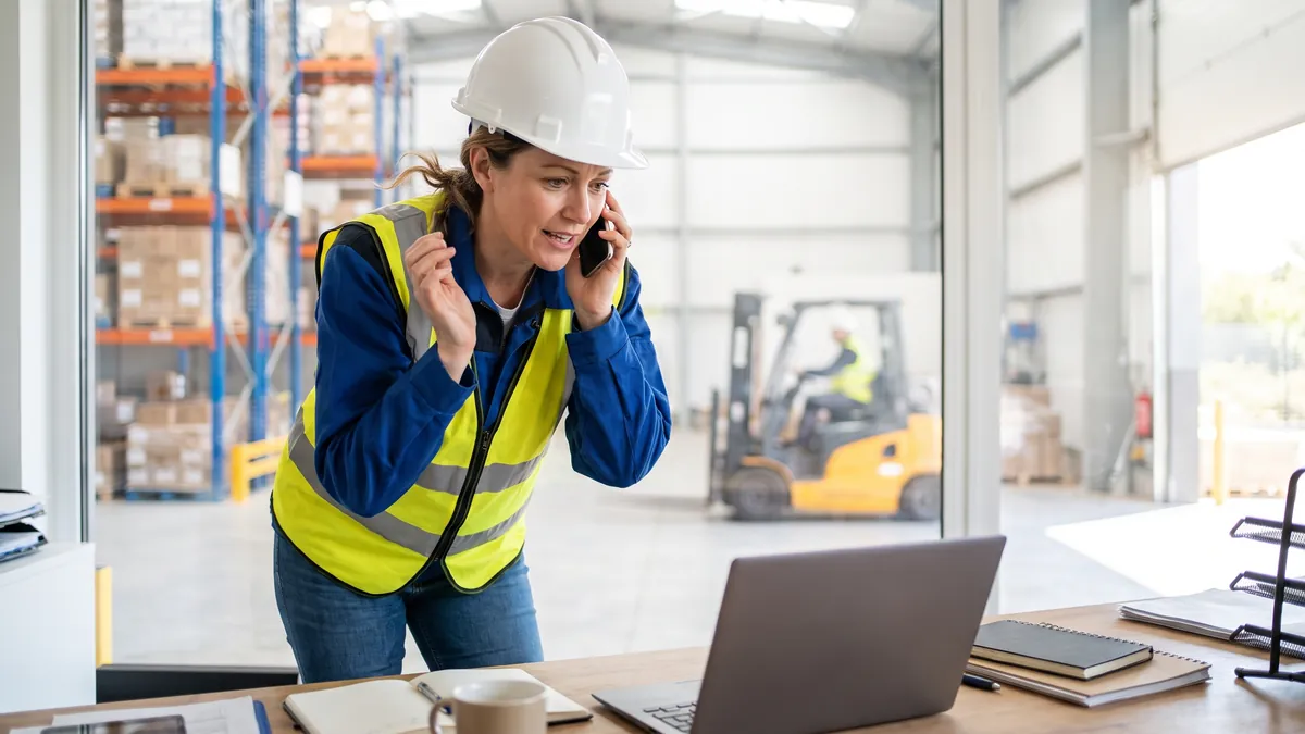 A woman in her forties in a hard hat and hi-vis vest talks on a phone at a bright warehouse office in Rotterdam, shelving and a forklift softly blurred behind her.