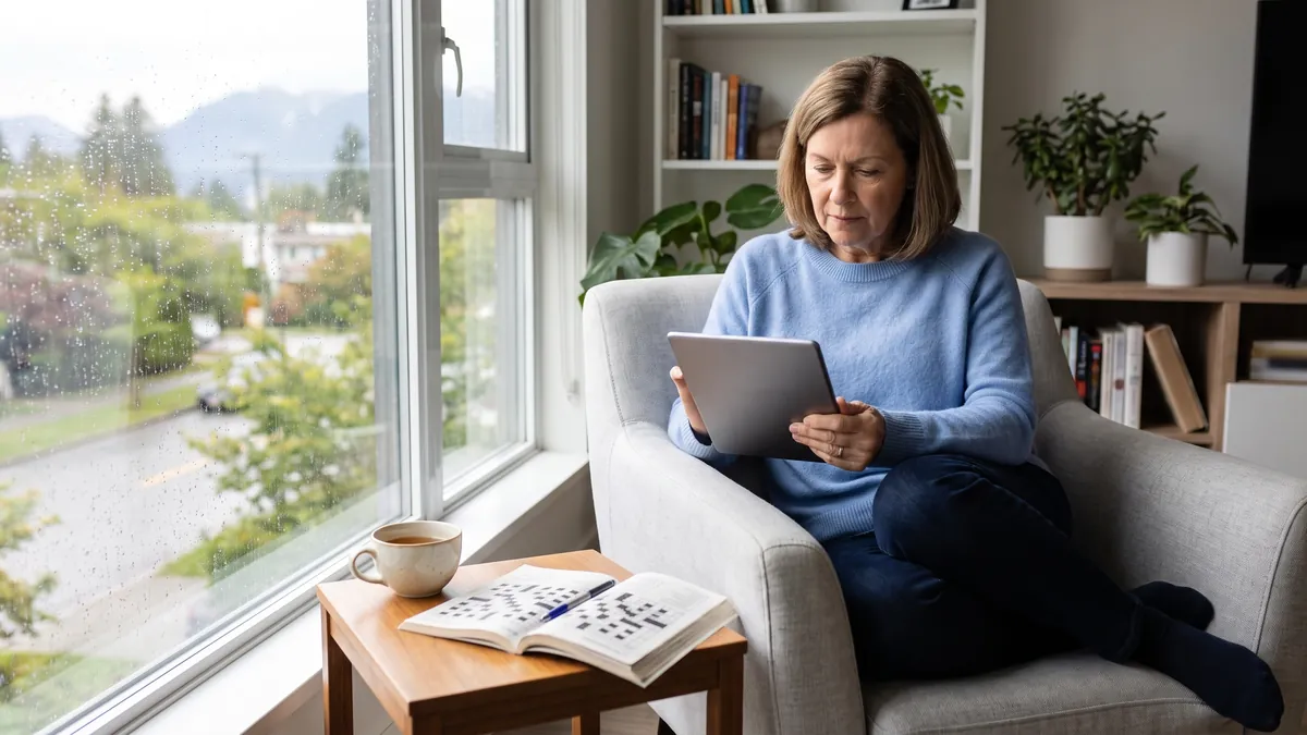 A woman in her sixties in Vancouver takes a survey on a tablet in a bright armchair by a window with a cup of tea and a crossword book.