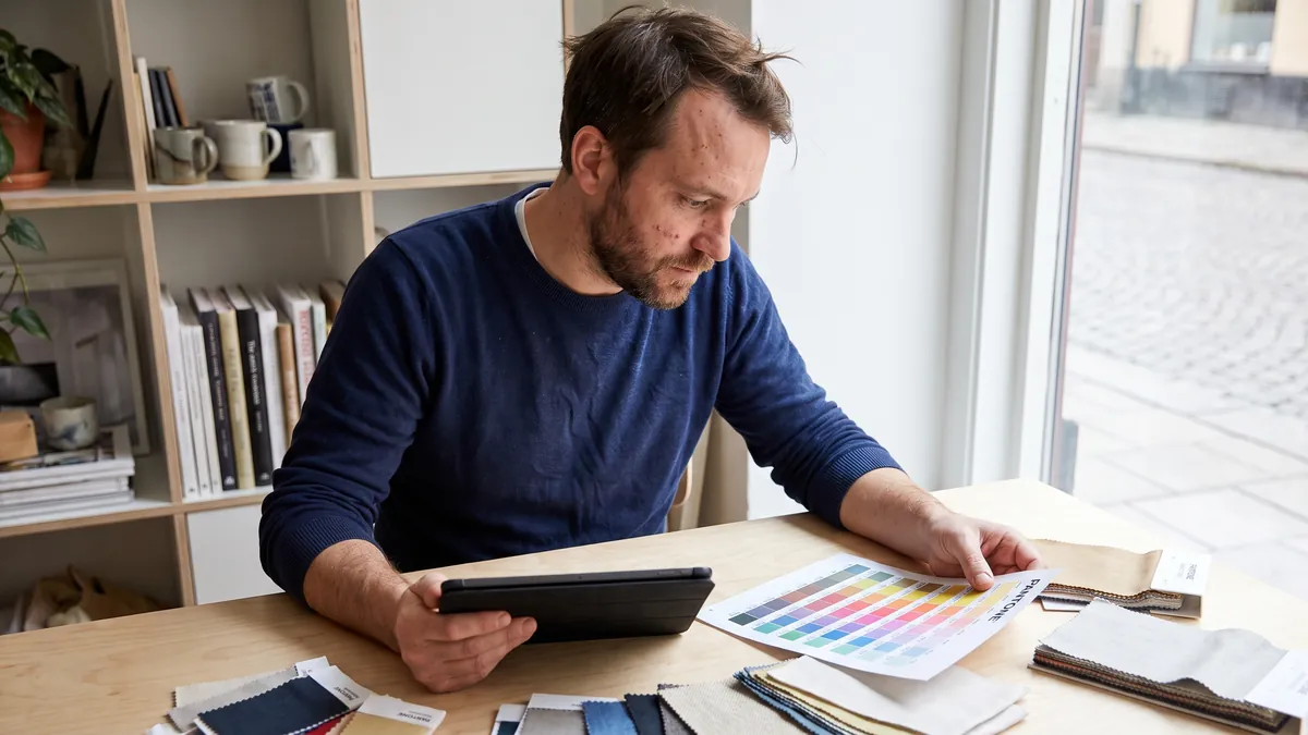 A man in his thirties in Stockholm looks at a printed colour chart while holding a tablet at a bright studio desk with design samples.