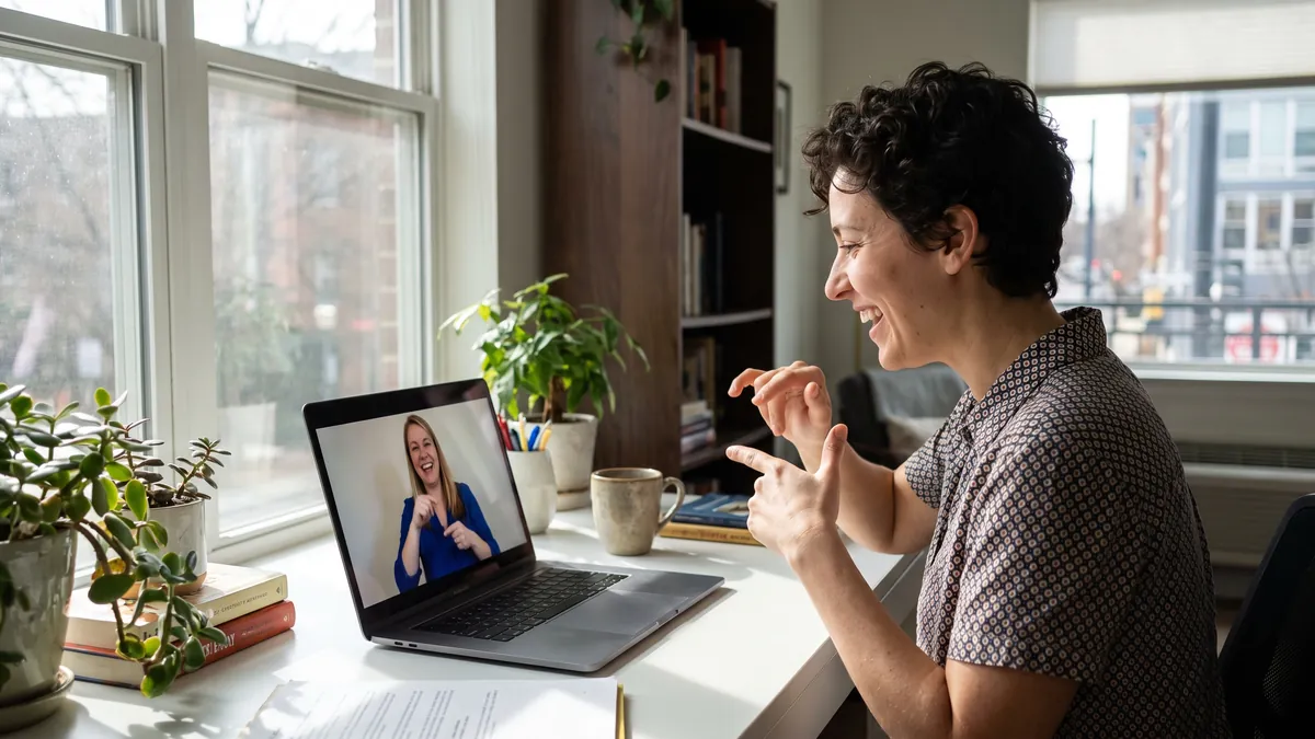 A woman in her thirties who is deaf signs to a sign language interpreter during a video session at a bright home office in Washington DC, both smiling.
