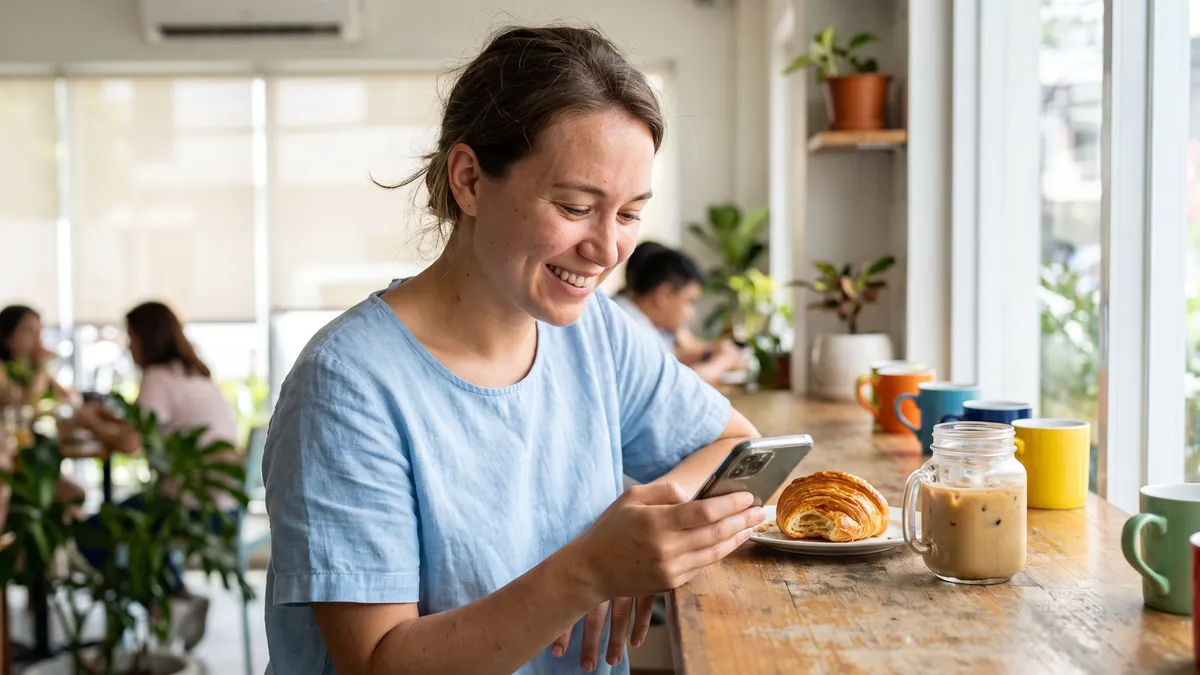 A woman in her twenties in Manila sits at a bright cafe counter looking at her phone and smiling, a pastry and an iced coffee beside her.
