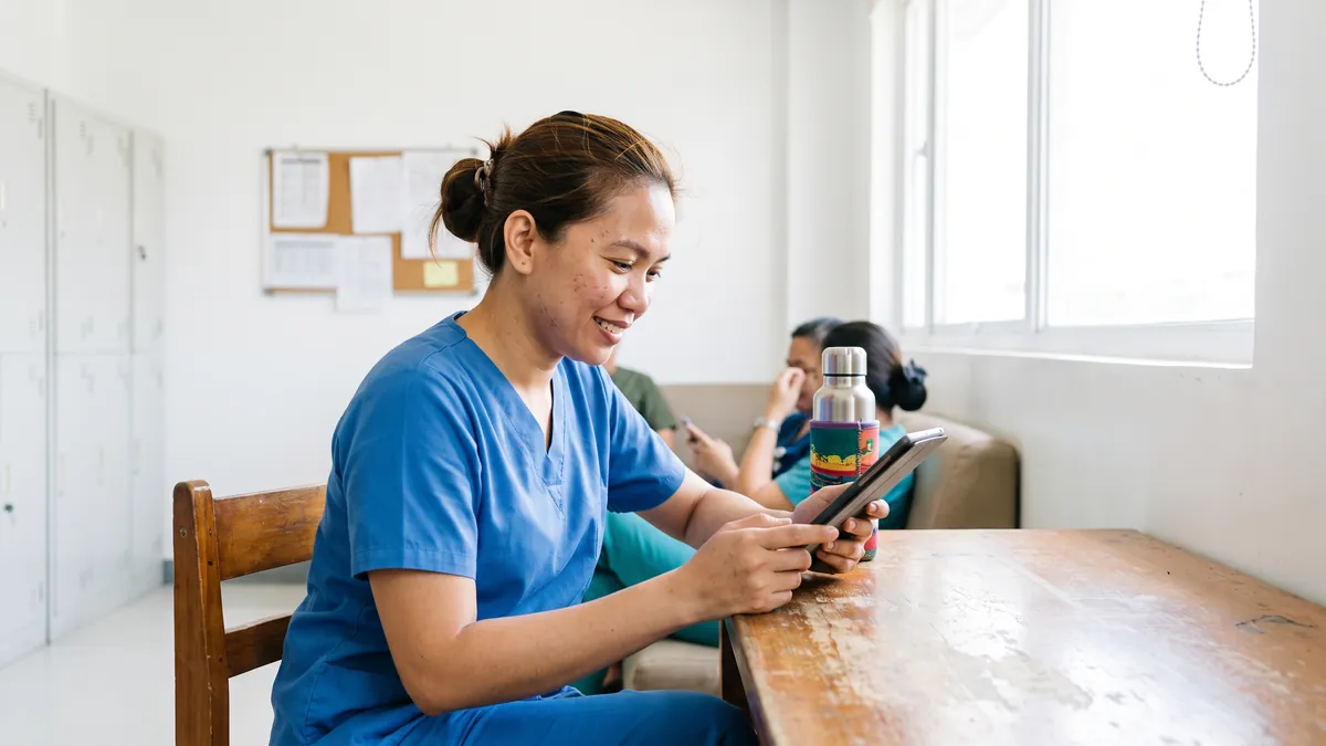 A nurse in her thirties in scrubs answers questions on a tablet during a break in a bright hospital staff room in Manila, a water bottle beside her.