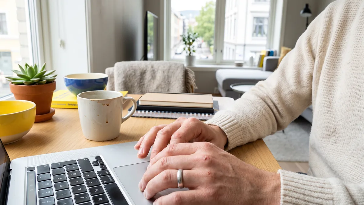 Close candid view of hands on a laptop keyboard at a bright desk in Oslo, a coffee mug and a small plant beside it, the screen out of frame.