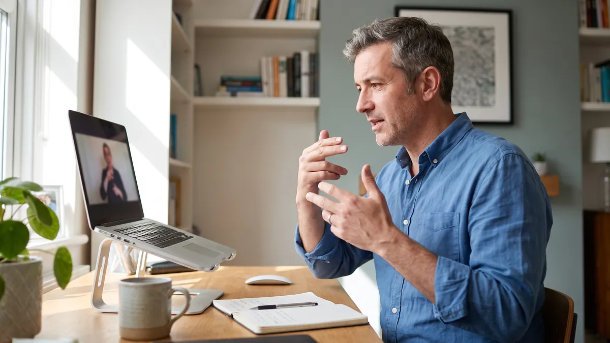 A deaf man in his forties signs during a bright online interview at a home desk in Leeds, an interpreter visible in a small window on the laptop screen, screen partly turned.
