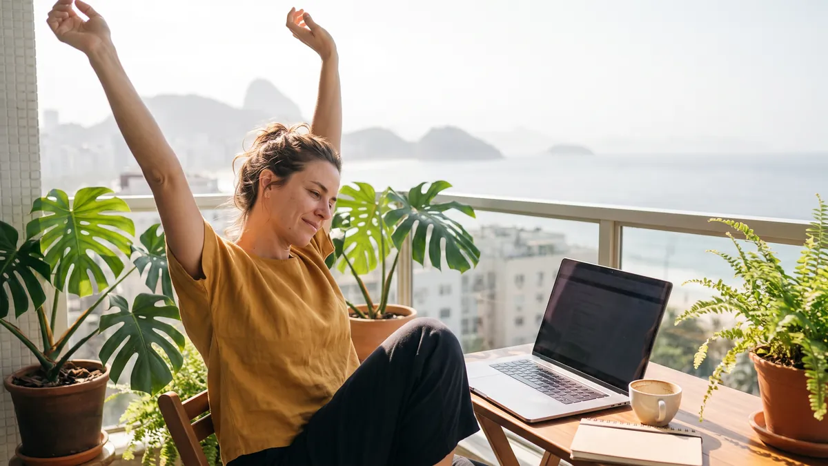 A woman in her thirties in Rio de Janeiro stretches on a bright balcony with plants after closing her laptop, ocean faintly visible in the distance.