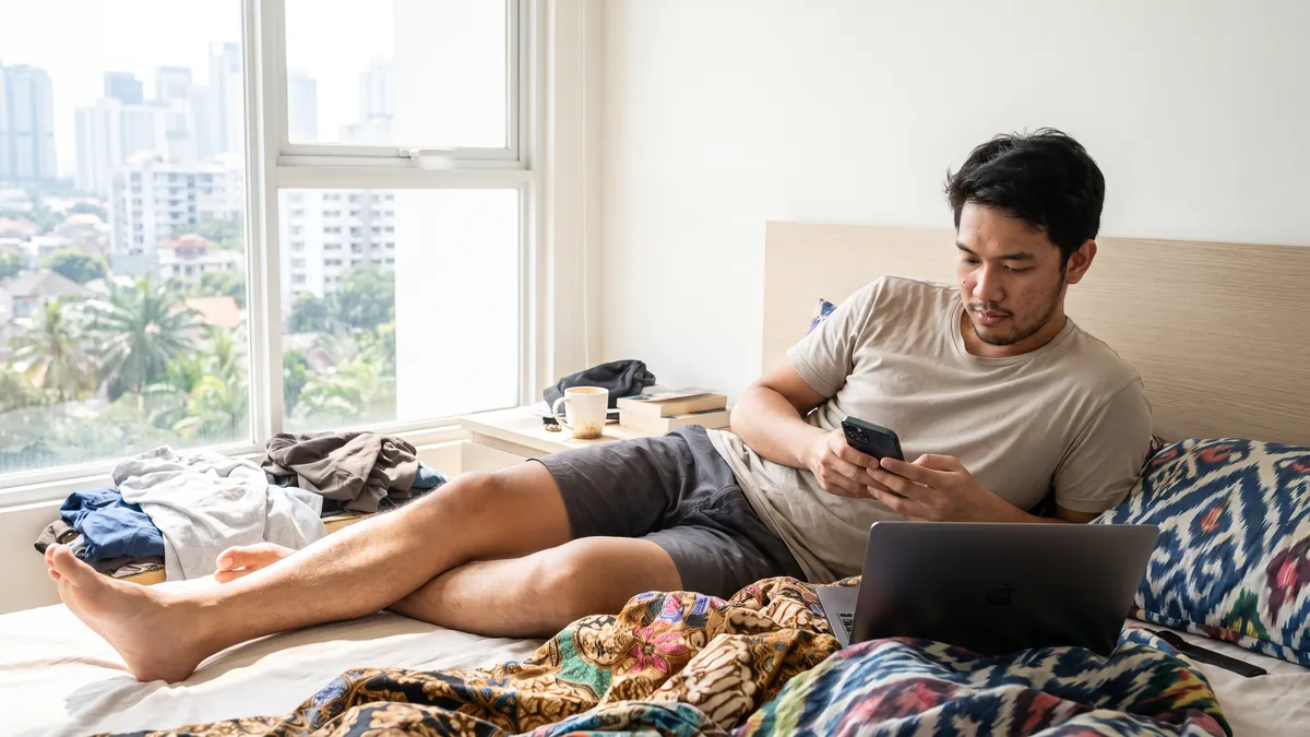 A man in his twenties in Jakarta lies on a bright bed with a phone in hand and a laptop open beside him, colourful bedding, late morning light.