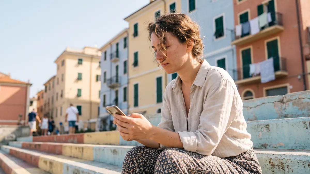 A woman in her twenties in Accra compares two things on her phone while sitting on bright outdoor steps, colourful buildings behind her.