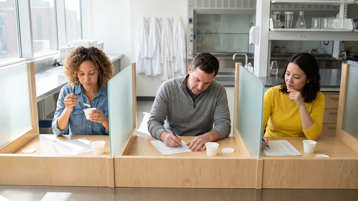 Three adults sit in bright tasting booths in a clean research facility in Chicago, tasting small unbranded cups of yogurt and marking paper forms.
