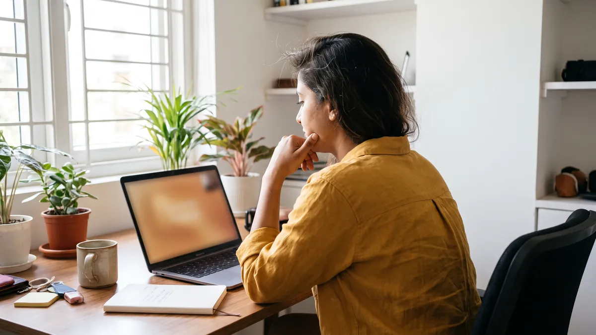 Over-the-shoulder view of a woman in Bengaluru on a video call at a home desk with plants and a window behind the laptop, the laptop screen softly out of focus and unreadable, warm daylight.