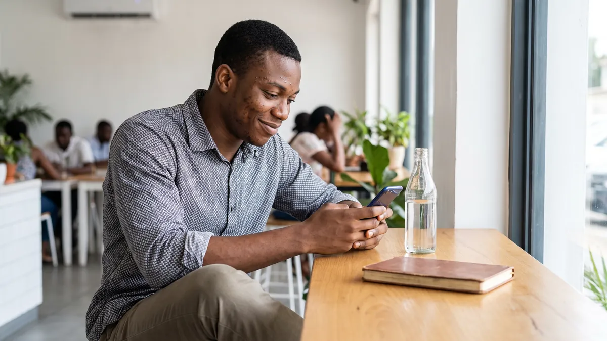 A young man in Lagos sits at a bright cafe table by the window looking at his phone with a slightly puzzled half smile, a bottle of water and a notebook on the table.